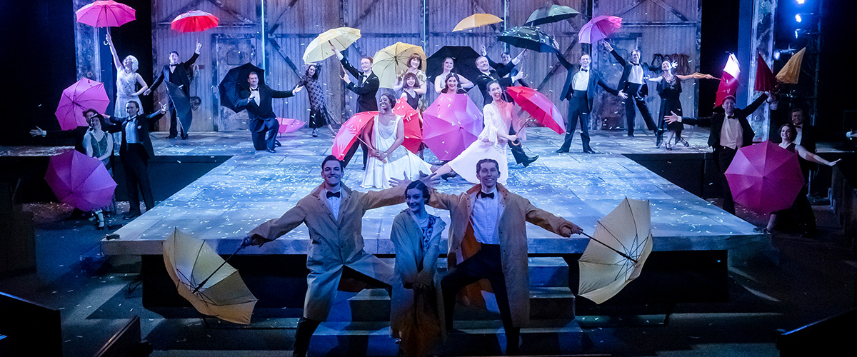 staged scene from Singin' in the Rain with cast on stage posed with umbrellas under blue light