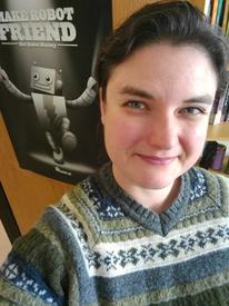 Smiling brunette white woman in fair isle sweater standing next to a poster that reads "Make Robot Friend, Not Robot Enemy"