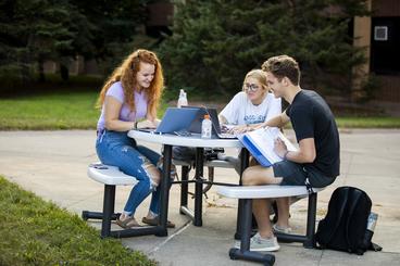 Three students laughing while sitting outside at a table with laptops.
